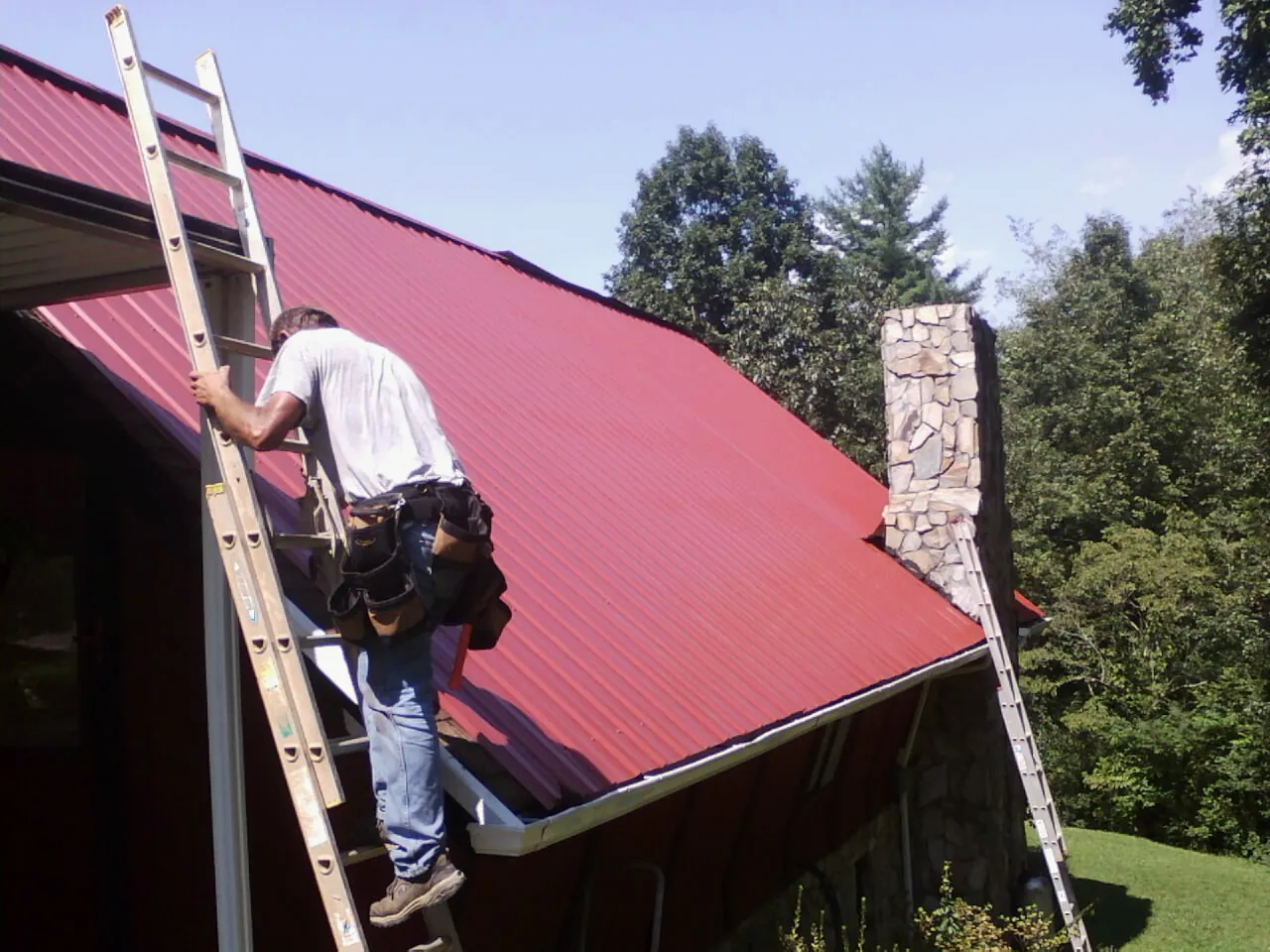 Roofer on a ladder beside a red metal roof and stone chimney — Soffit & Fascia Repair by Roofing Brothers in Cerulean